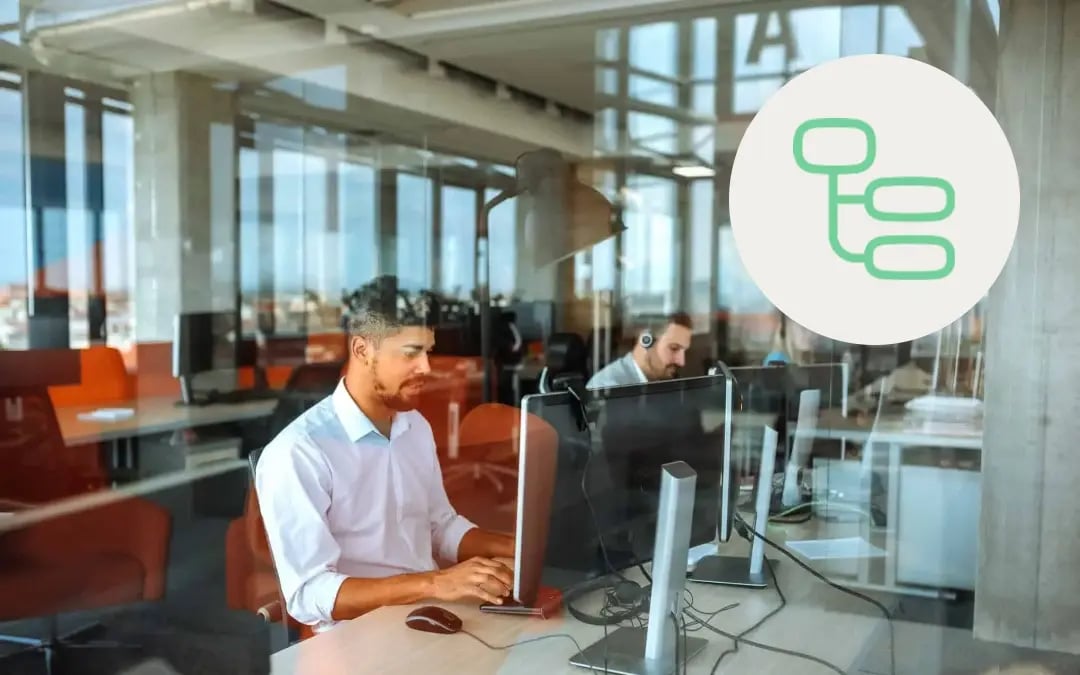 A contact center agent sitting at a desk in front of a computer in a modern office setting with large glass windows, with a green data icon overlaid on the image.