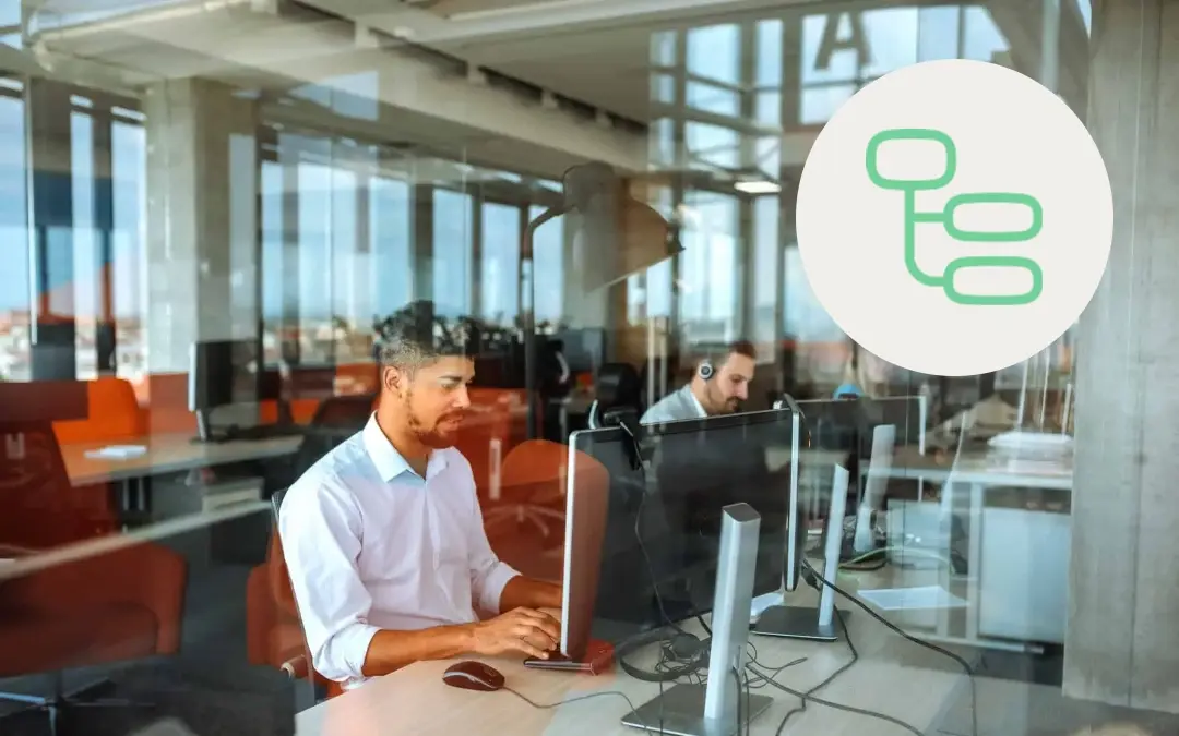 A contact center agent sitting at a desk in front of a computer in a modern office setting with large glass windows, with a green data icon overlaid on the image.