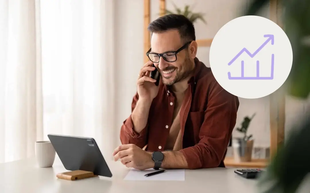 Smiling man in glasses on a phone call at his desk, looking at a tablet, next to a purple icon of a rising trend line over a bar chart